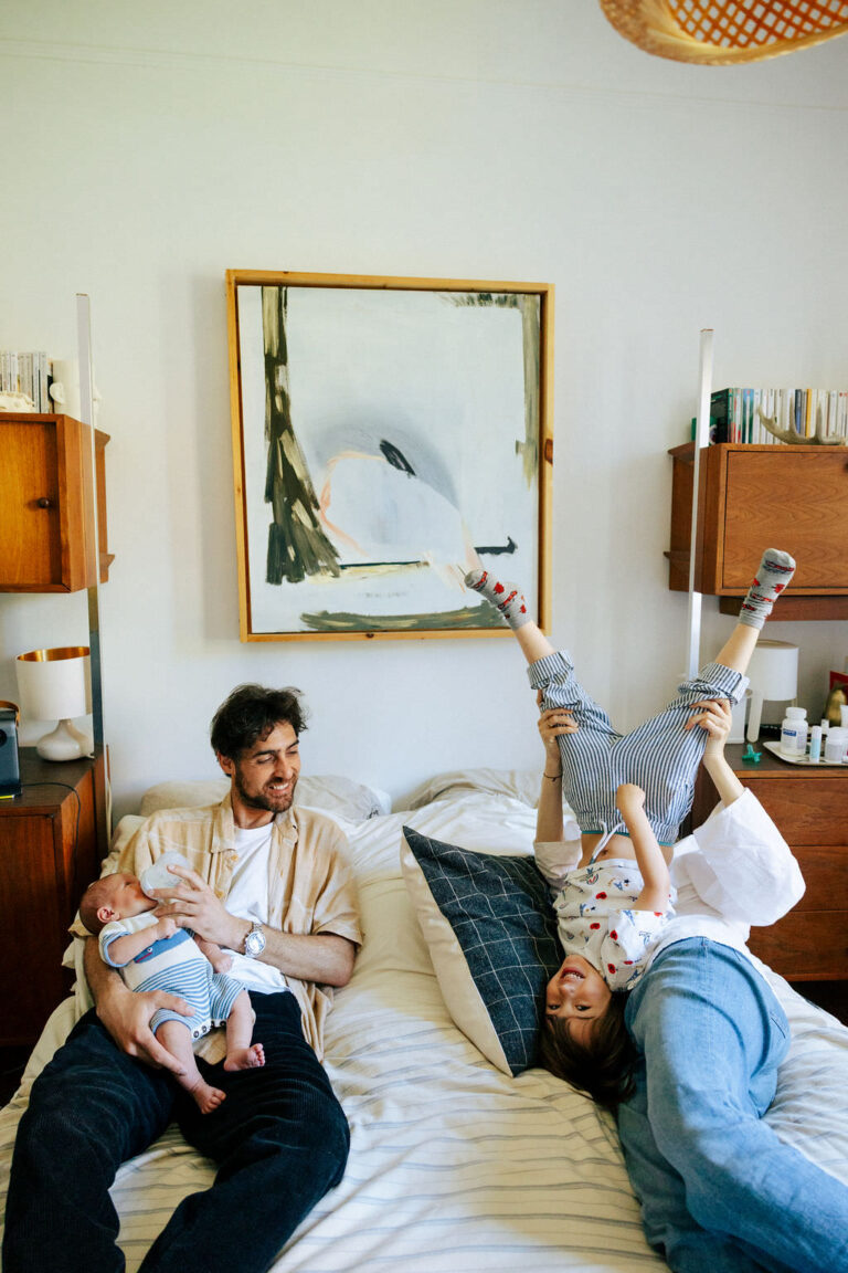 A man sits on a bed holding a baby, while two children play beside him; one child is upside down with legs in the air, and the other is lying across the bed. Captured by an NYC family photographer in a cozy, modern bedroom.