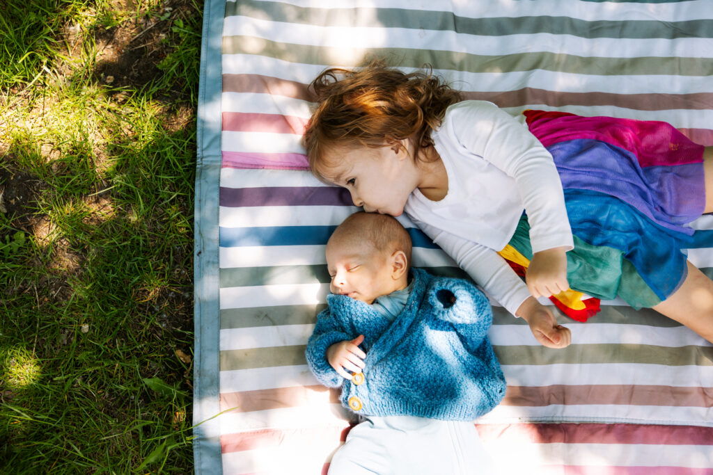 A young child in a colorful skirt lies on a striped blanket outdoors, gently cuddling and kissing a baby dressed in a blue sweater—capturing the warmth and joy of outdoor newborn photography NYC, with sunlight and grass at the edge of the scene.