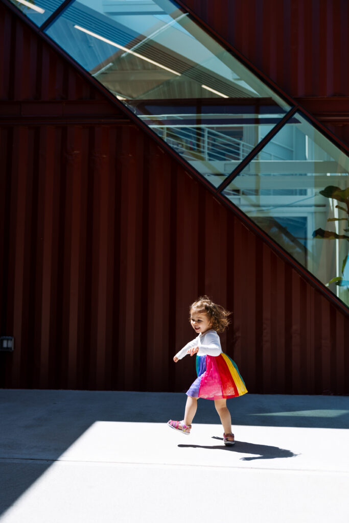 A young child wearing a colorful rainbow dress walks in sunlight in front of a modern building with red walls and geometric glass windows, capturing the vibrant spirit of outdoor newborn photography NYC.
