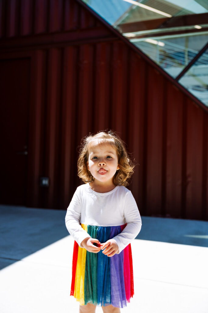 A young child with light brown hair wears a rainbow-colored dress and stands outdoors on a sunny day in front of a modern red building with large windows, capturing the vibrant spirit often seen in outdoor newborn photography NYC.