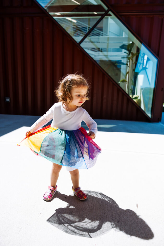 A young child in a white shirt and rainbow-colored skirt stands outdoors in sunlight, holding the sides of her skirt. Perfect for outdoor newborn photography NYC, she casts a shadow as a modern building with large windows rises behind her.