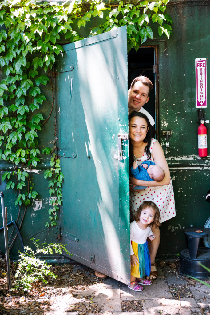 A smiling family of four stands in a doorway surrounded by green ivy. Capturing an Outdoor newborn photography NYC moment, an adult man and woman holding a baby and a young girl peek out from behind a sunlit green door.