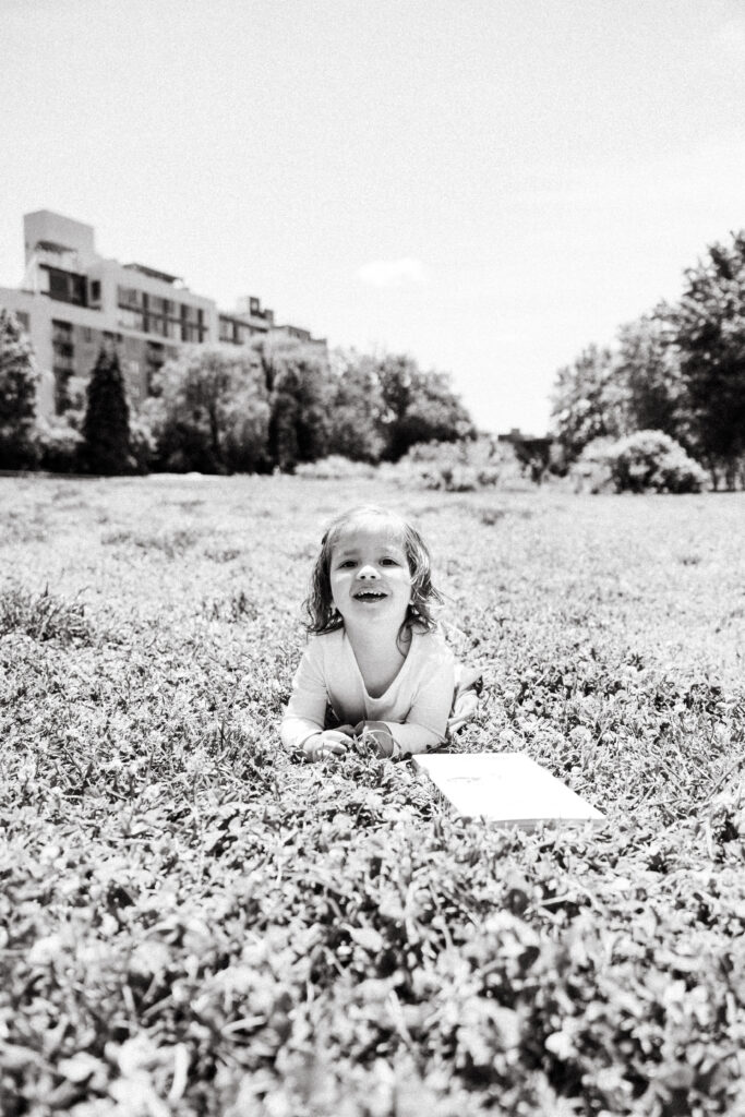 A young child lies on their stomach in a grassy field, smiling and looking up. In front of the child is an open book or notebook. This charming black and white photo captures the essence of outdoor newborn photography NYC. Trees and buildings frame the bright sky.
