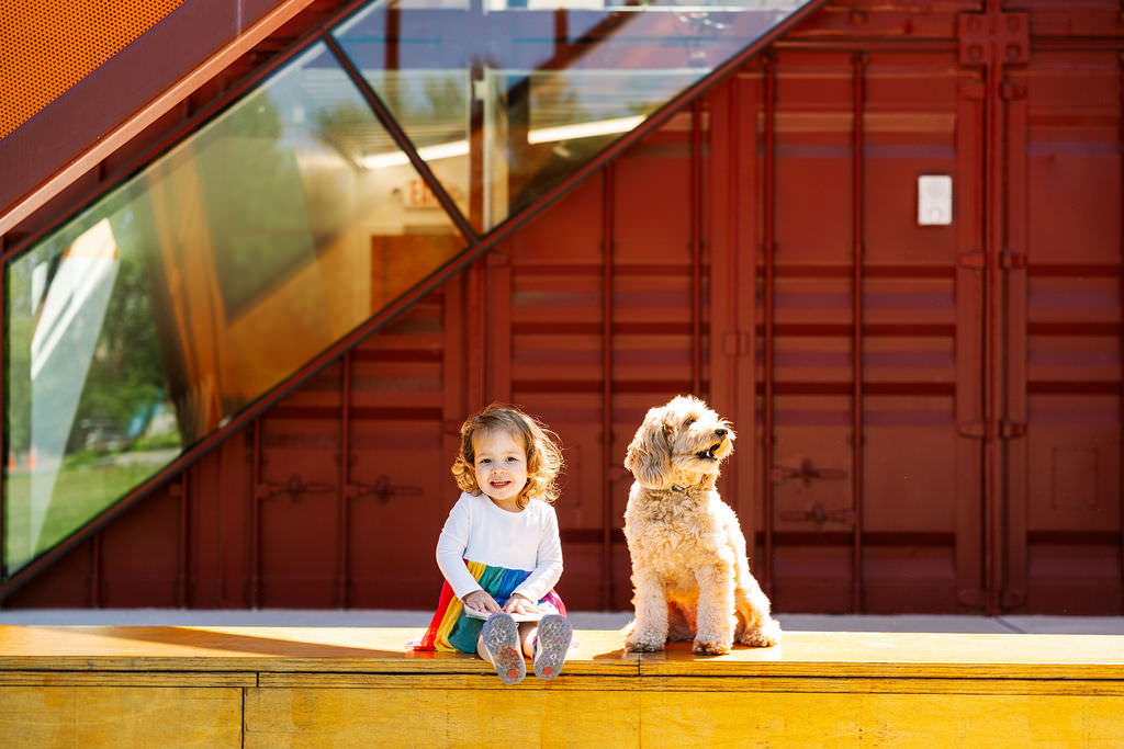 A young girl in a colorful dress and cape sits smiling next to a fluffy dog on a yellow platform, with a red, modern building featuring large glass windows behind them—capturing the charm of outdoor newborn photography NYC style.