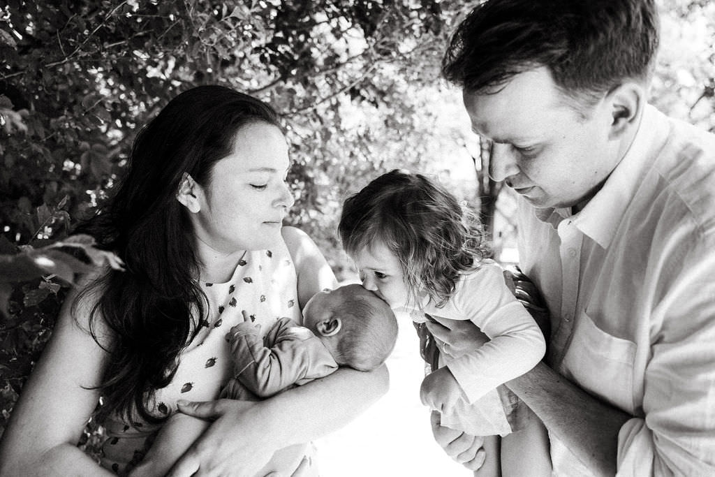Black and white portrait of toddler sister gently kissing newborn baby brother during family session