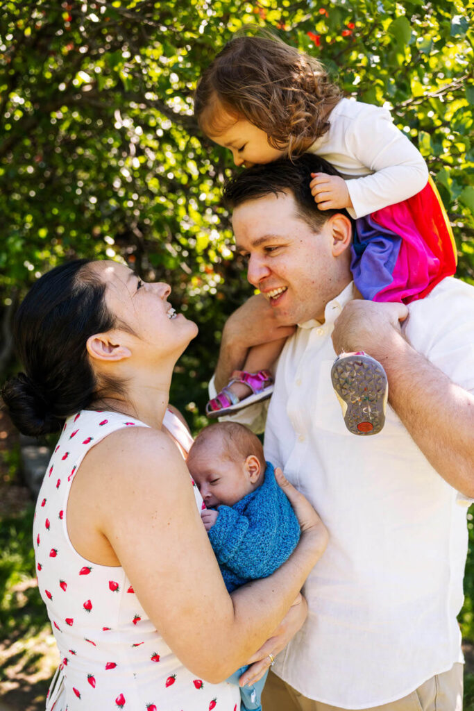 A smiling family of four stands outdoors in a sunlit park. The mother cradles a sleeping baby in a blue blanket, showcasing outdoor newborn photography NYC, while the father carries a toddler on his shoulders as sunlight filters through leafy trees.