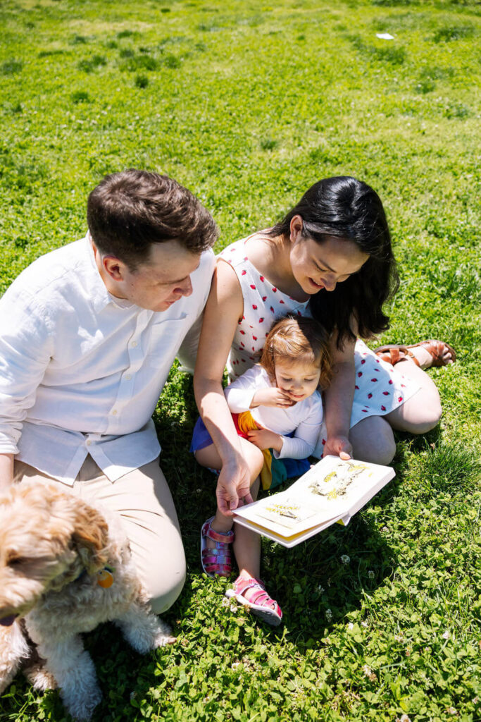 A family sits on green grass outdoors. An adult man, woman, and young child read a book together while their small dog sits nearby. The sun is shining, creating a perfect scene for outdoor newborn photography NYC, with everyone looking engaged and happy.
