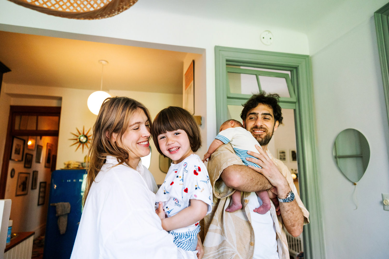 A happy family stands together in a bright room. A smiling woman holds a cheerful young child, while a man cradles a baby in his arms—capturing the warmth and joy of home, perfect for newborn photography.
